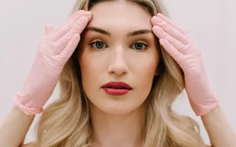 Kobieta z blond włosami w różowych rękawiczkach dotyka skroni. Przygotowanie do zabiegu, jakim jest liposukcja twarzy, by uzyskać idealny kontur.